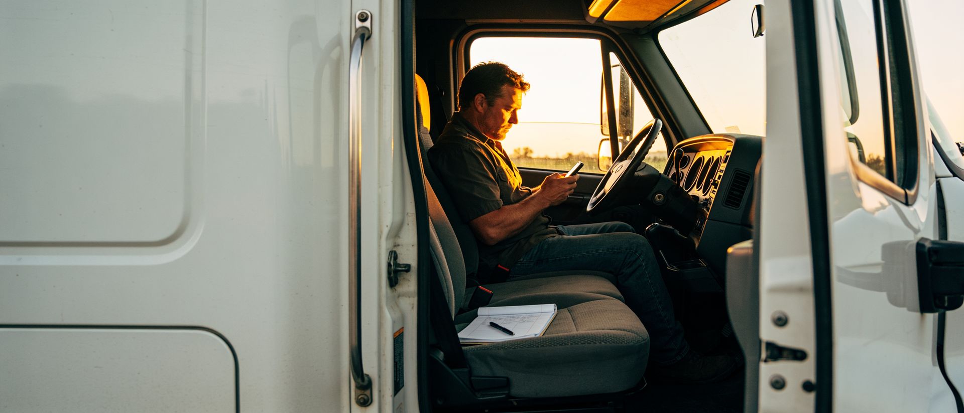 Driver in the cab of his truck at dusk, looking at his phone, logbook on the seat beside him